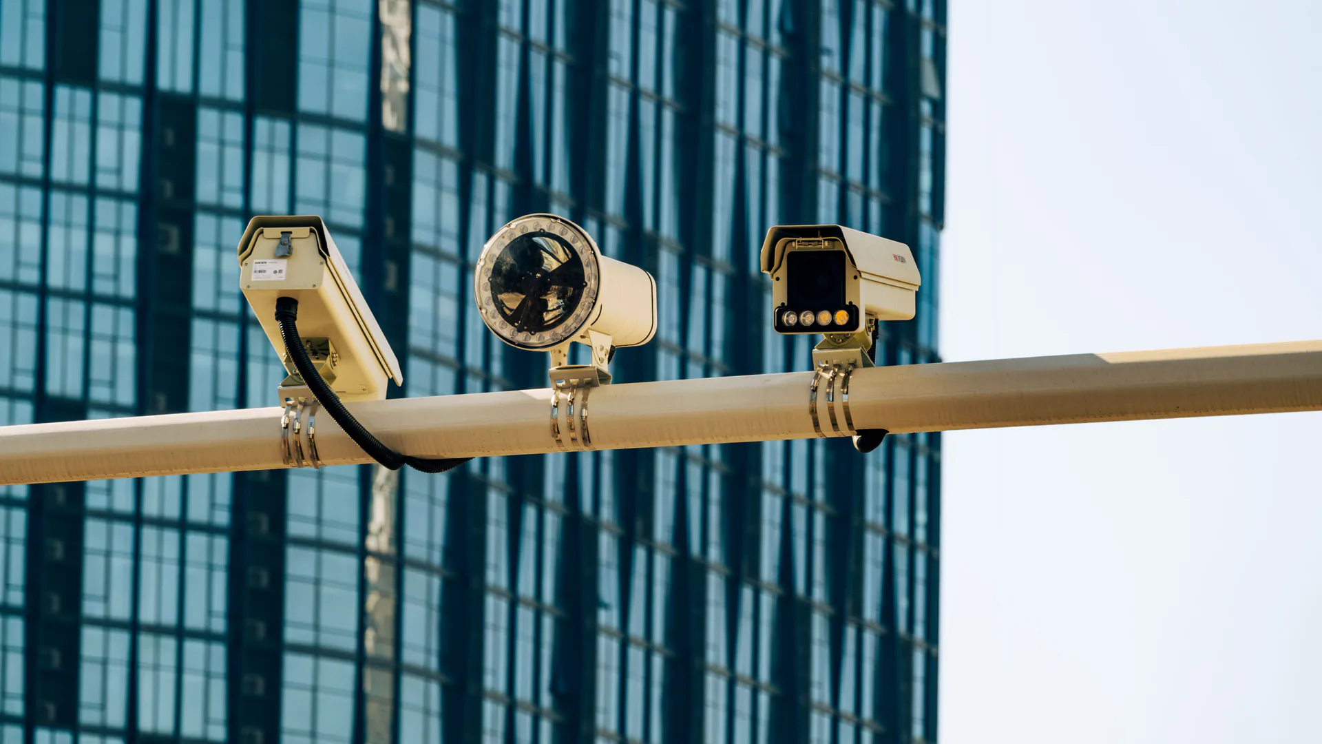 Several fixed security cameras mounted on a horizontal support outside modern buildings