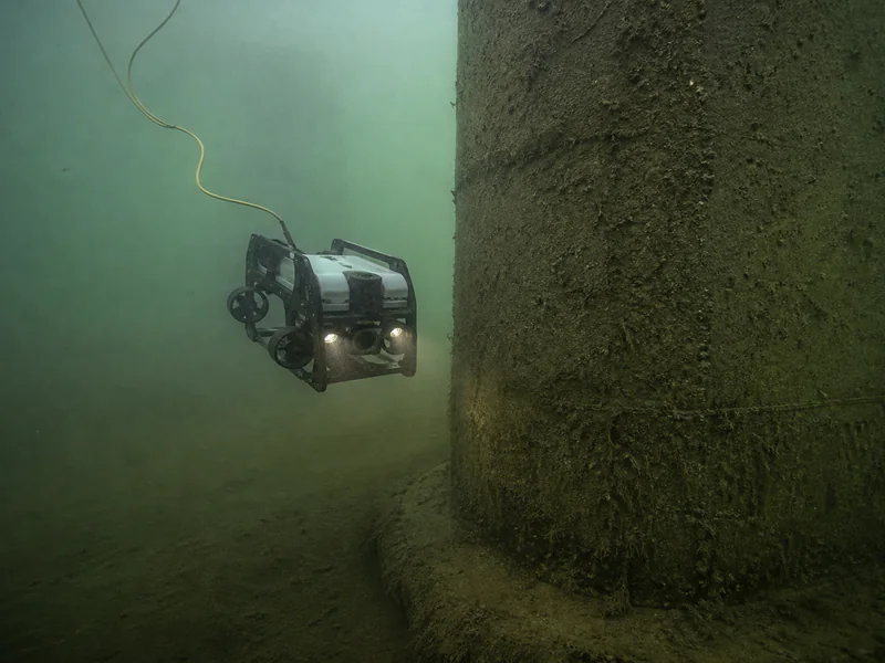 Tethered inspection ROV imaging an algae-covered concrete bridge pier in naturally turbid river water